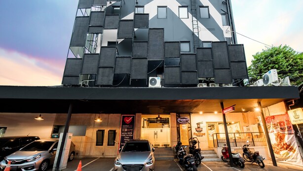 En moderne bygning med en geometrisk facade, en tydelig indgang og en parkeringsplads med biler og motorcykler.