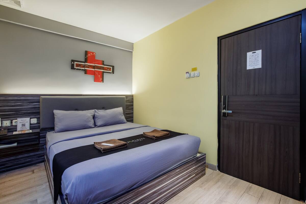 A modern hotel room with a large bed, a headboard with a built-in desk, a wall-mounted artwork, and a dark wooden door.