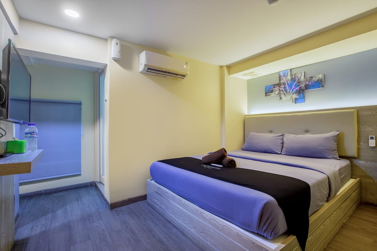 A hotel room with a bed, a small table with a water bottle, and an air conditioning unit on the wall.