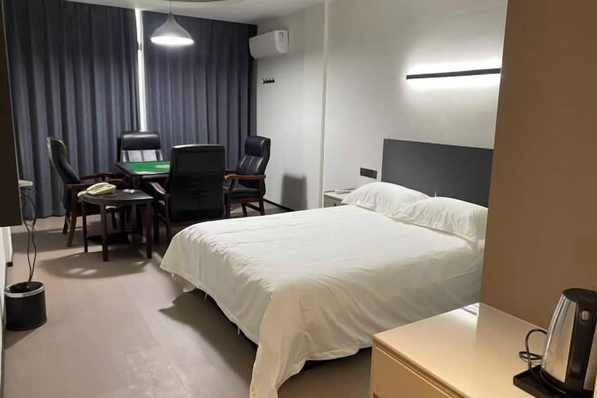 A hotel room with a bed, a desk with a chair, a telephone, and a bedside table with a kettle.