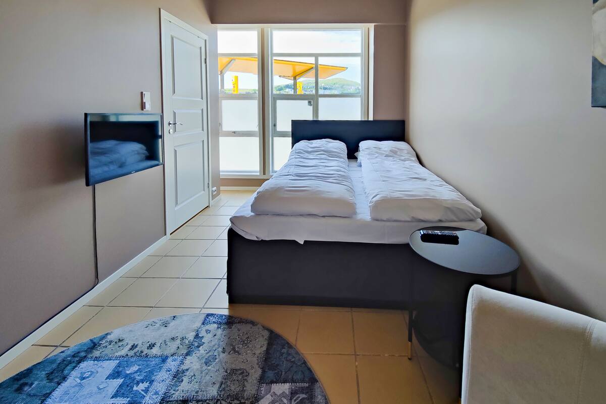 A narrow hotel room with a bed, a TV mounted on the wall, a small table, and a view of the outside through a window.