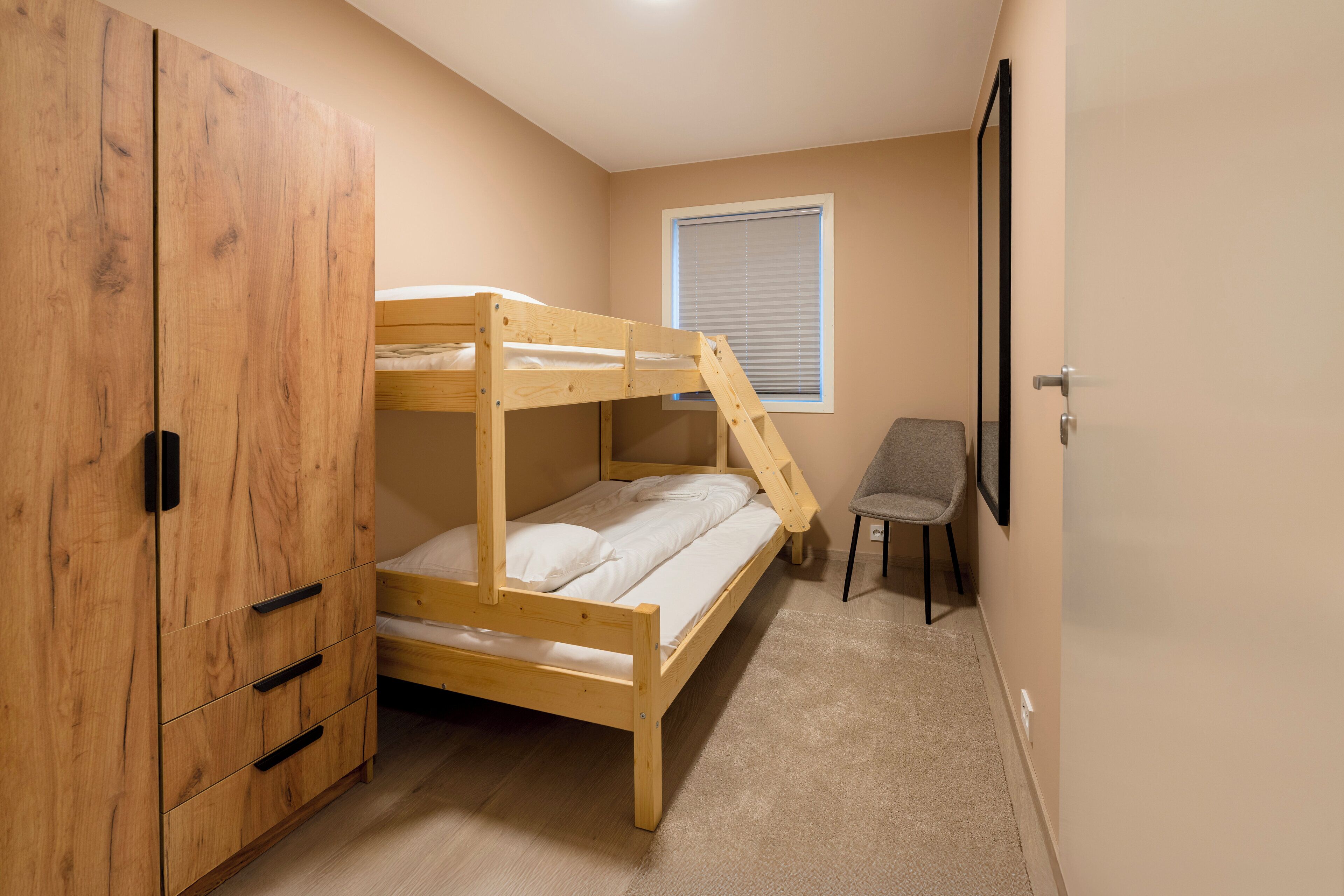 A bunk bed with a wooden frame, a wardrobe with multiple drawers, a window with blinds, and a chair.