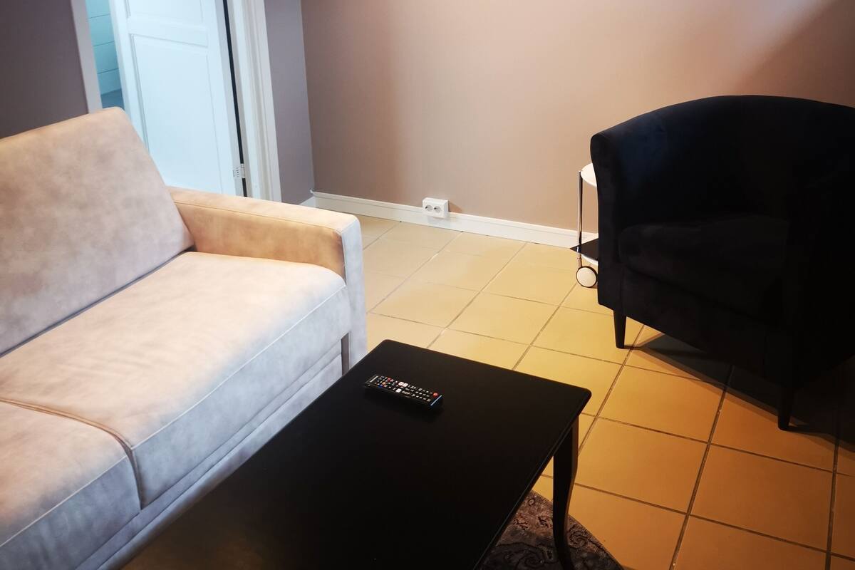 A living room with a beige sofa, a black coffee table, and a dark armchair. A white door is visible in the background.