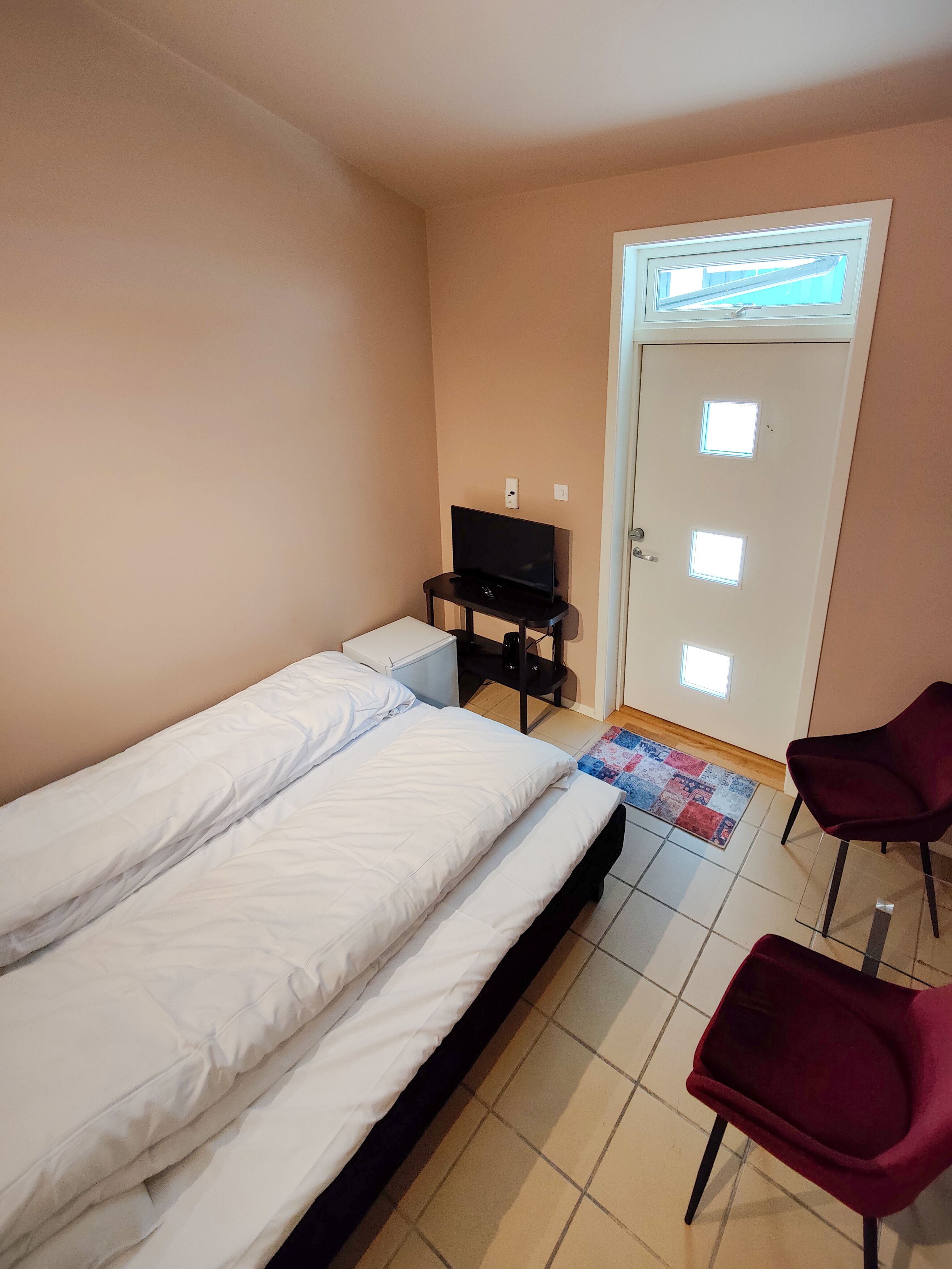 Quarto com duas camas de solteiro, uma TV sobre um suporte, uma porta com janelas e duas cadeiras.