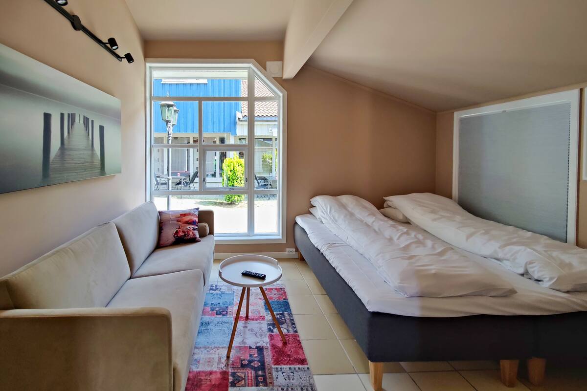A compact hotel room with a bed, a sofa, a small table, and a large window offering a view of the outside.