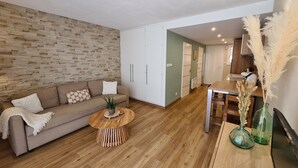 A modern living room with a wooden floor, a brown sofa, a round coffee table, and a kitchen area visible through an open door. - Appartement au Calme Dans Residence Avec Piscine, Entouré de Foret (Bœrsch)
