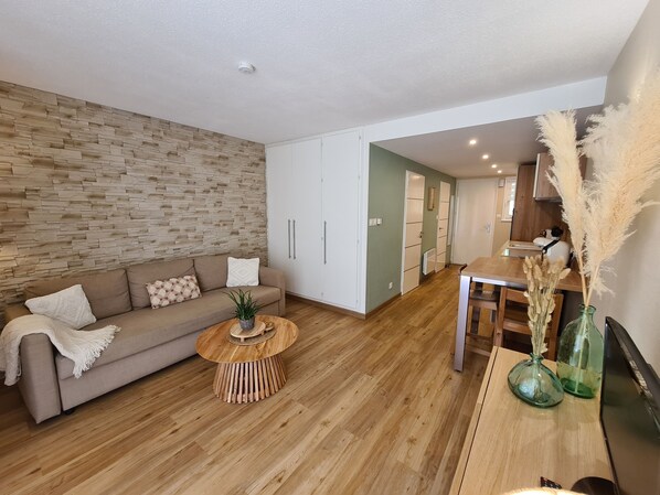 Uma sala de estar moderna com piso de madeira, um sofá marrom, uma mesa de centro redonda e uma área de cozinha visível através de uma porta aberta.