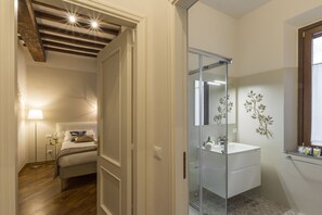 A hotel room with a bed, bedside lamp, and a bathroom with a glass shower enclosure and a sink. - Casa Dora by Mmega (Florence)