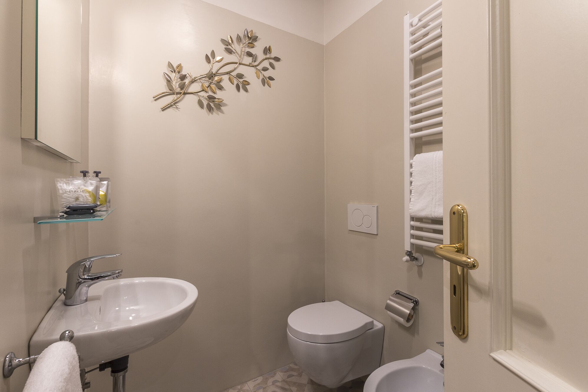 A bathroom with a white sink, toilet, and bathtub. There is a towel rack with a towel, a decorative wall piece, and a towel warmer.
