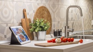 Una cocina moderna con un lavamanos de acero inoxidable, una tableta sobre un soporte, una planta en maceta y tomates frescos sobre una tabla de cortar.