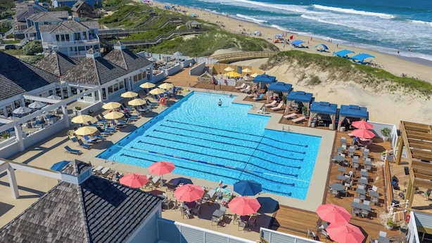 Piscina com espreguiçadeiras e guarda-sóis, praia com pessoas e vista para o mar.