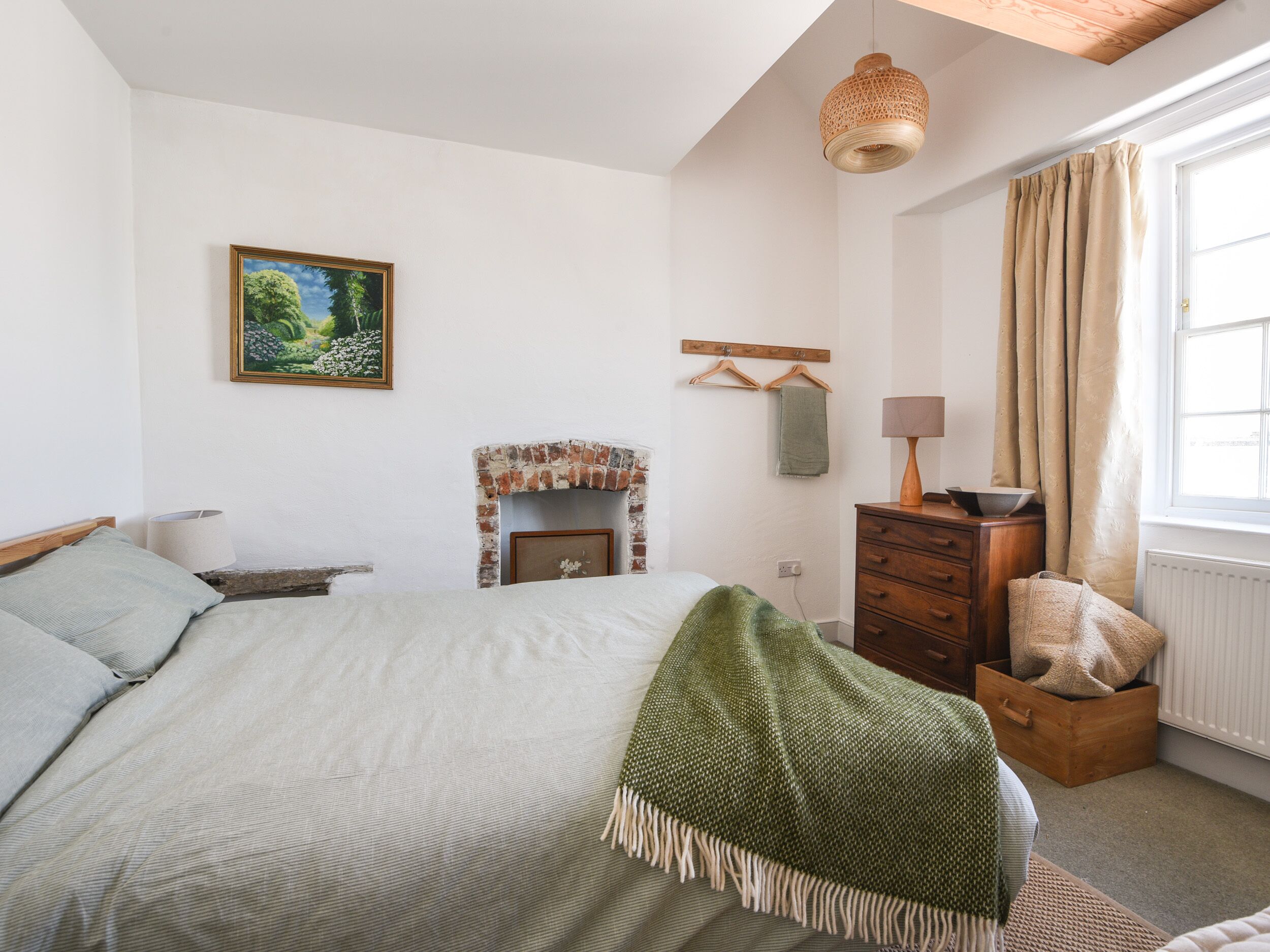 A bedroom with a bed, a fireplace, a wooden dresser, a hanging lamp, and a window with curtains.