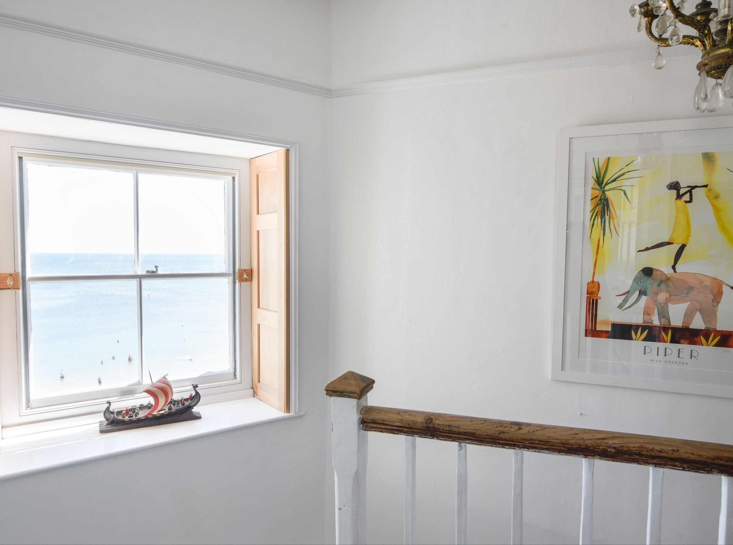A room with a large window overlooking the sea, a small ship model on the windowsill, and a framed artwork on the wall.