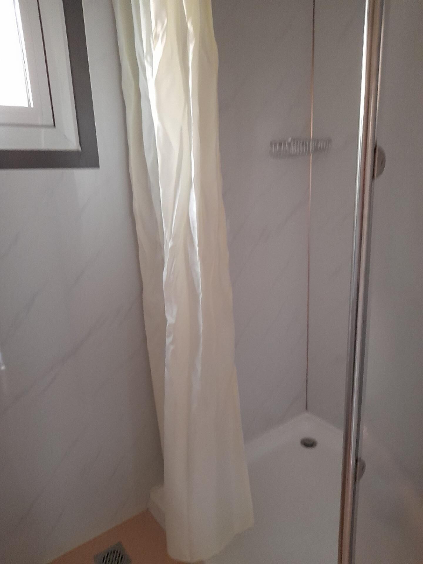 Un área de ducha pequeña y cerrada con una cortina blanca, un cabezal de ducha metálico y un suelo de baldosas.