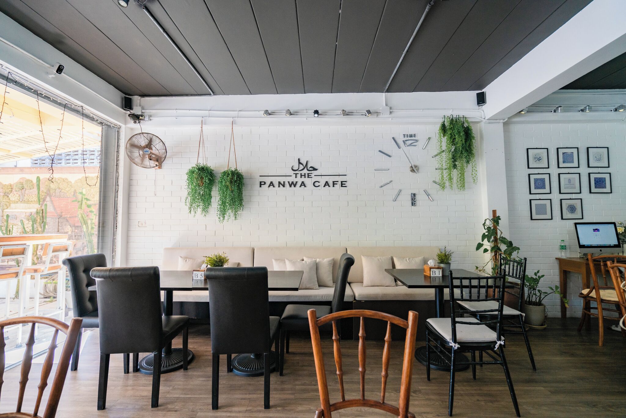 L’intérieur d’un café avec un mur en briques blanches, un canapé, des chaises noires et en bois, et des plantes suspendues.