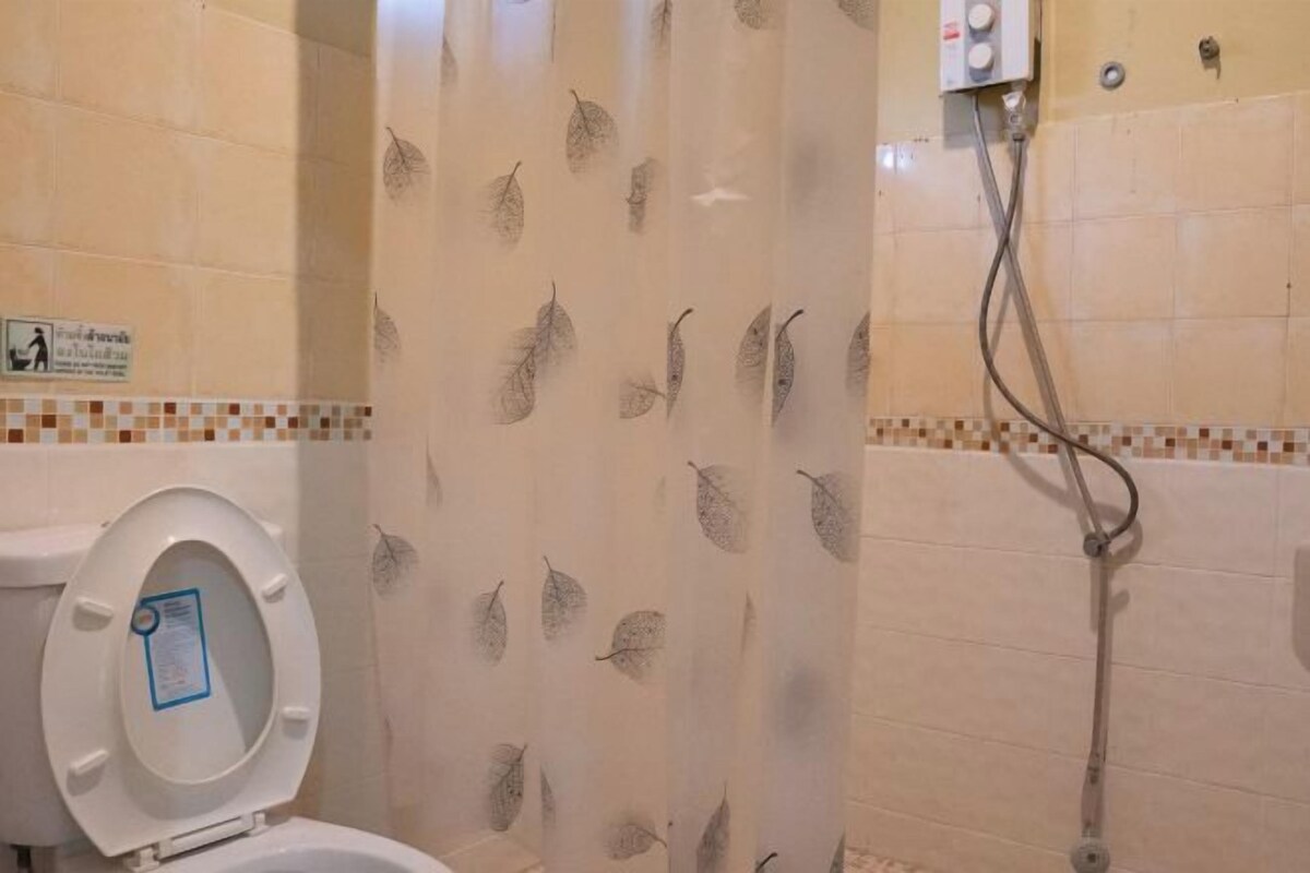 A bathroom with a toilet, a shower with a leaf-patterned shower curtain, and a tiled wall with a small sign.