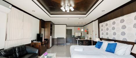 A modern hotel room with a large bed, a black leather sofa, a small coffee table, and a kitchen area in the background.