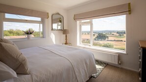 A bedroom with a large bed, a window with a view of the countryside, a bedside table with a lamp, and a mirror on the wall. - Ridgeway Farmhouse at Ragley Estate, Alcester - sleeps 8 guests  in 4 bedrooms (Alcester)