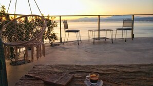 Une terrasse avec un fauteuil suspendu, deux chaises, une table avec une tasse de café et une vue sur la mer et les montagnes.