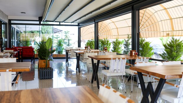 Restaurante moderno com mesas de madeira e cadeiras brancas, grandes janelas e plantas em vasos.