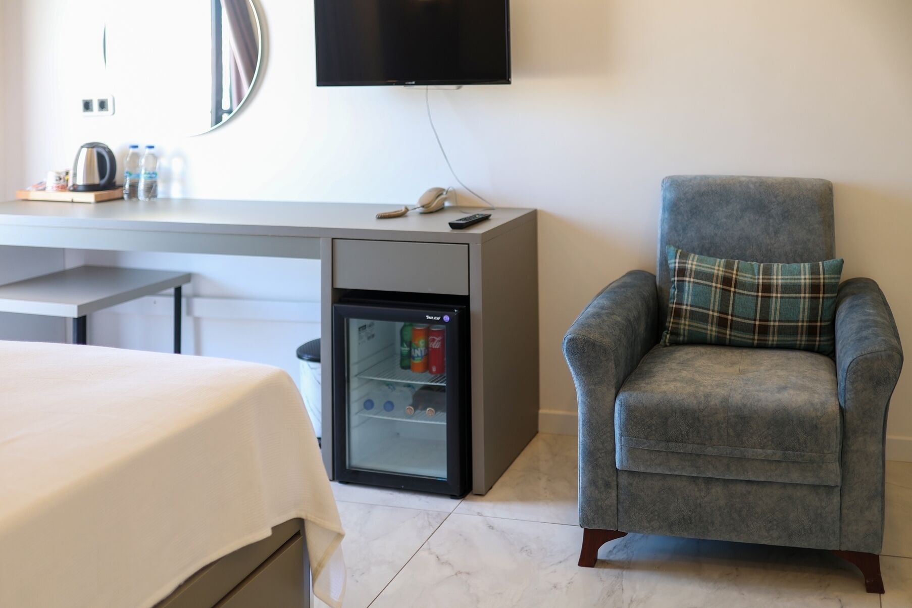 A hotel room with a bed, a desk with a refrigerator, a chair with a plaid cushion, and a flat-screen TV mounted on the wall.