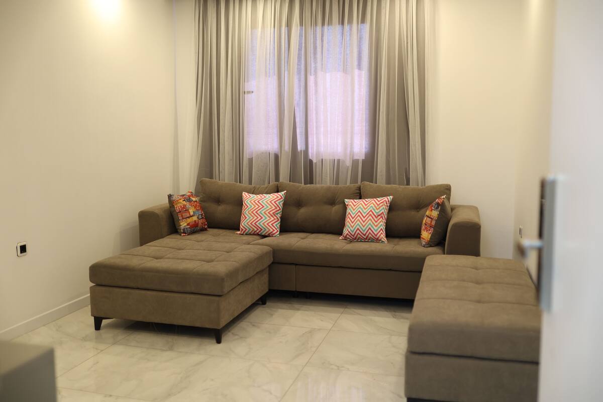 Un salon moderne avec un canapé d’angle, un pouf et des coussins à motifs.