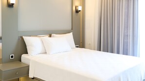 Cama bem arrumada com lençóis brancos, mesa de cabeceira com abajur e vista para uma janela com cortinas.