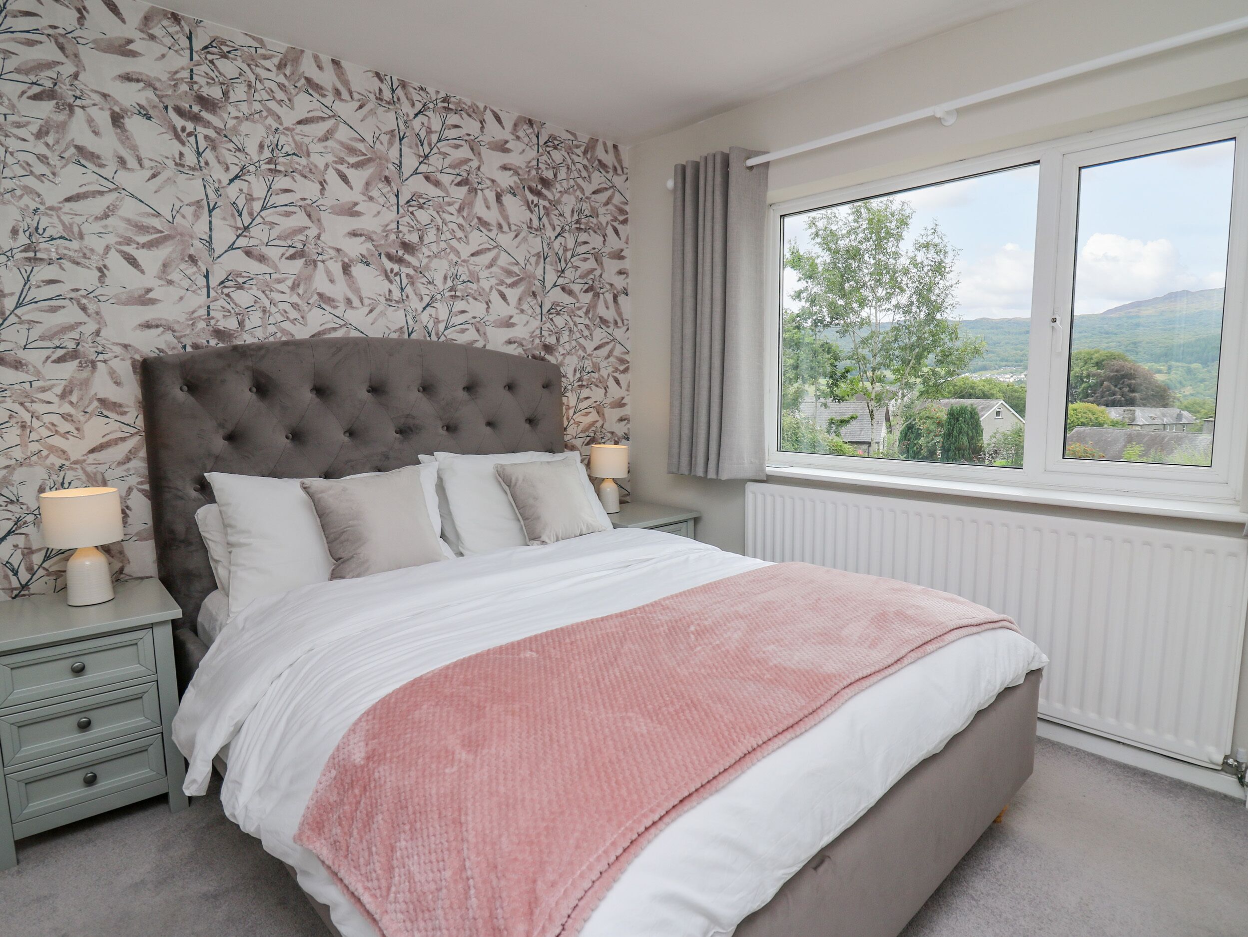 A bedroom with a large bed, a headboard, two bedside tables with lamps, a window with a view, and a floral wallpaper.