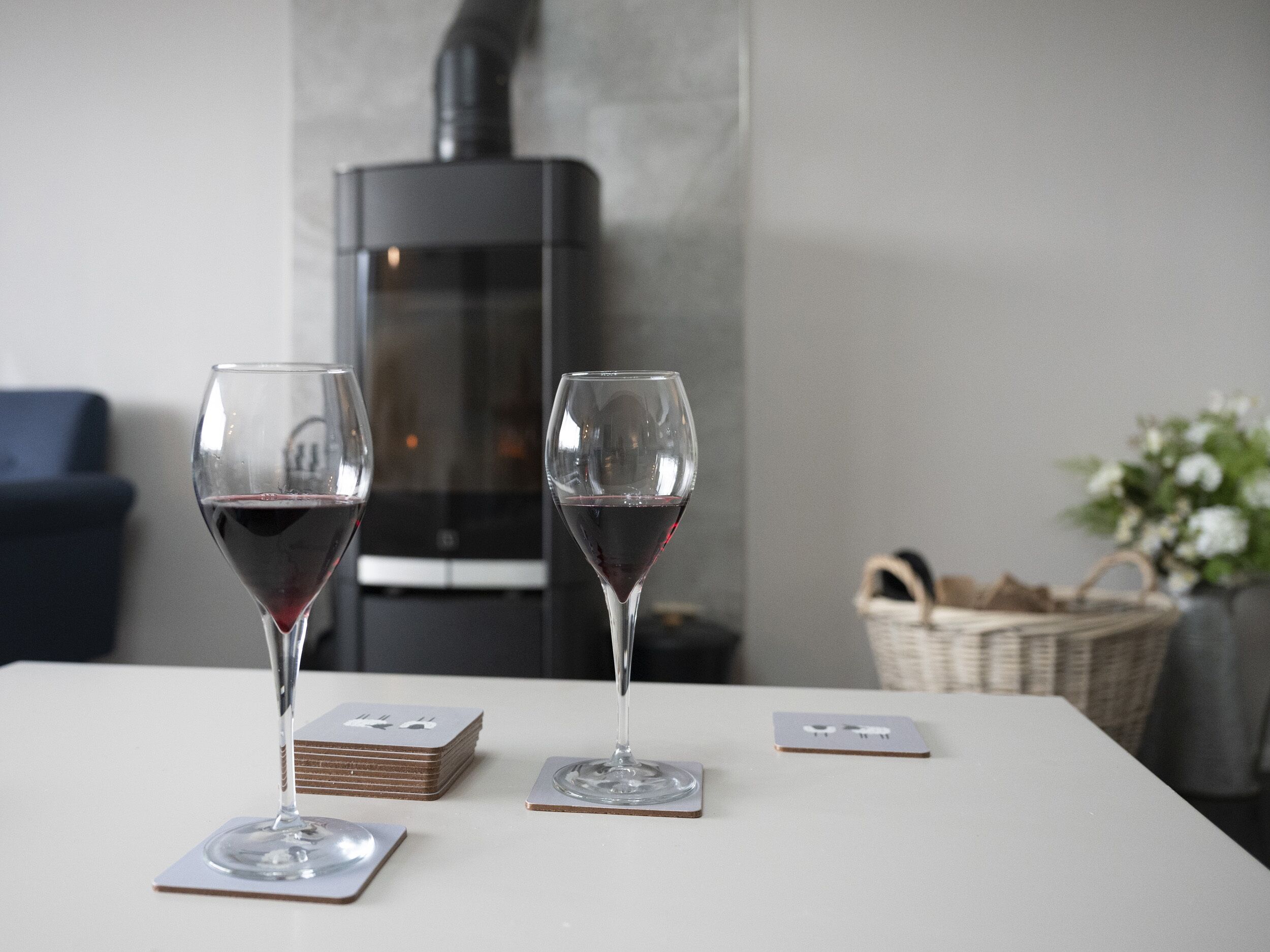 Two wine glasses on a table with coaster sets, a fireplace, and a wicker basket in the background.