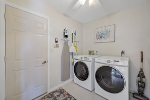A laundry room with two front-loading washing machines, a wall-mounted ironing station, a vacuum cleaner, and a door.