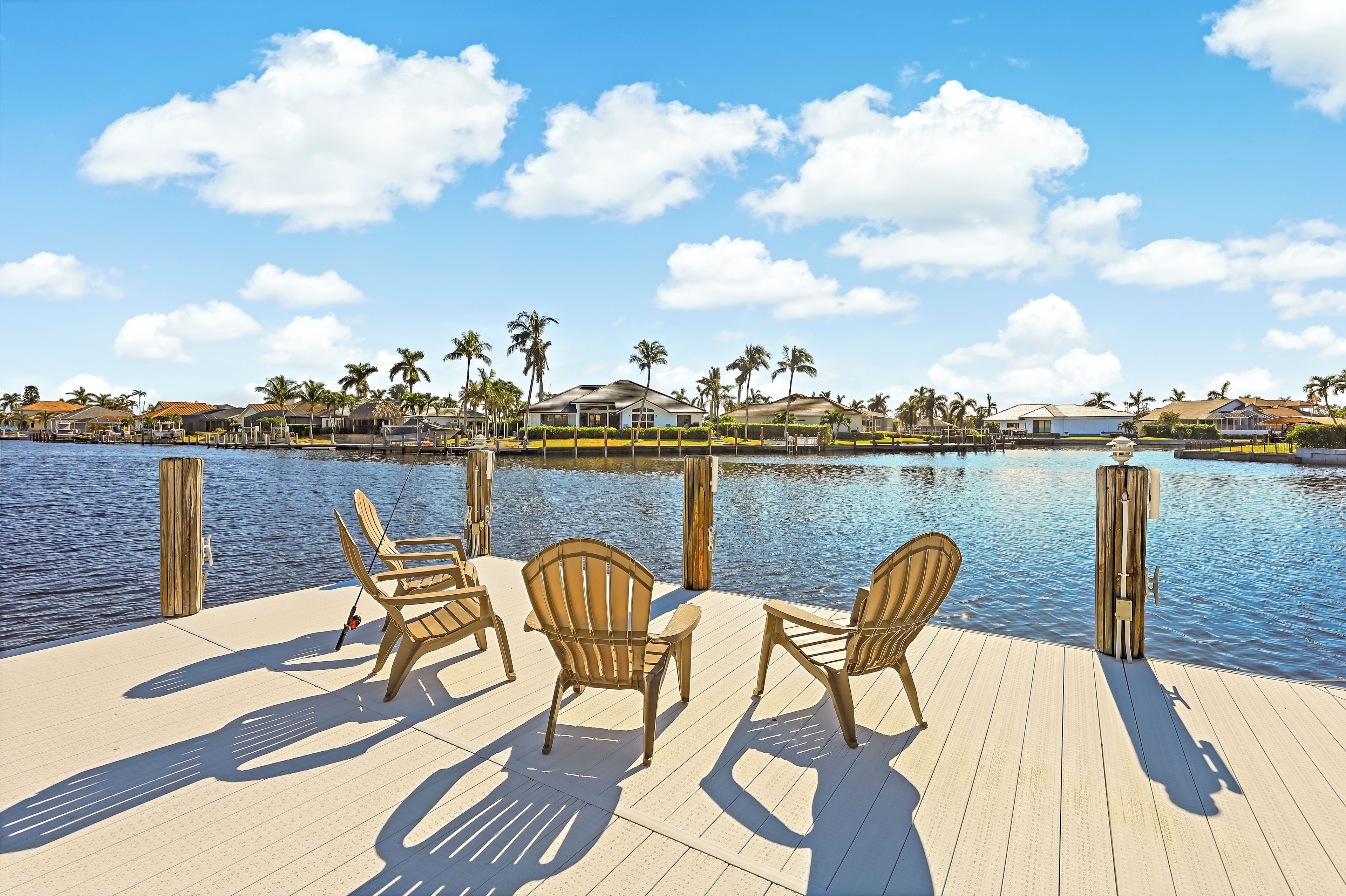 Une terrasse en bois, agrémentée de chaises, qui offre une vue sur une étendue d’eau calme, entourée de maisons et de palmiers.
