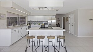 A modern kitchen with white cabinetry, a central island with stools, and a large window. - Direct Boating Access Home | Pool/Spa & Billiards (Marco Island)