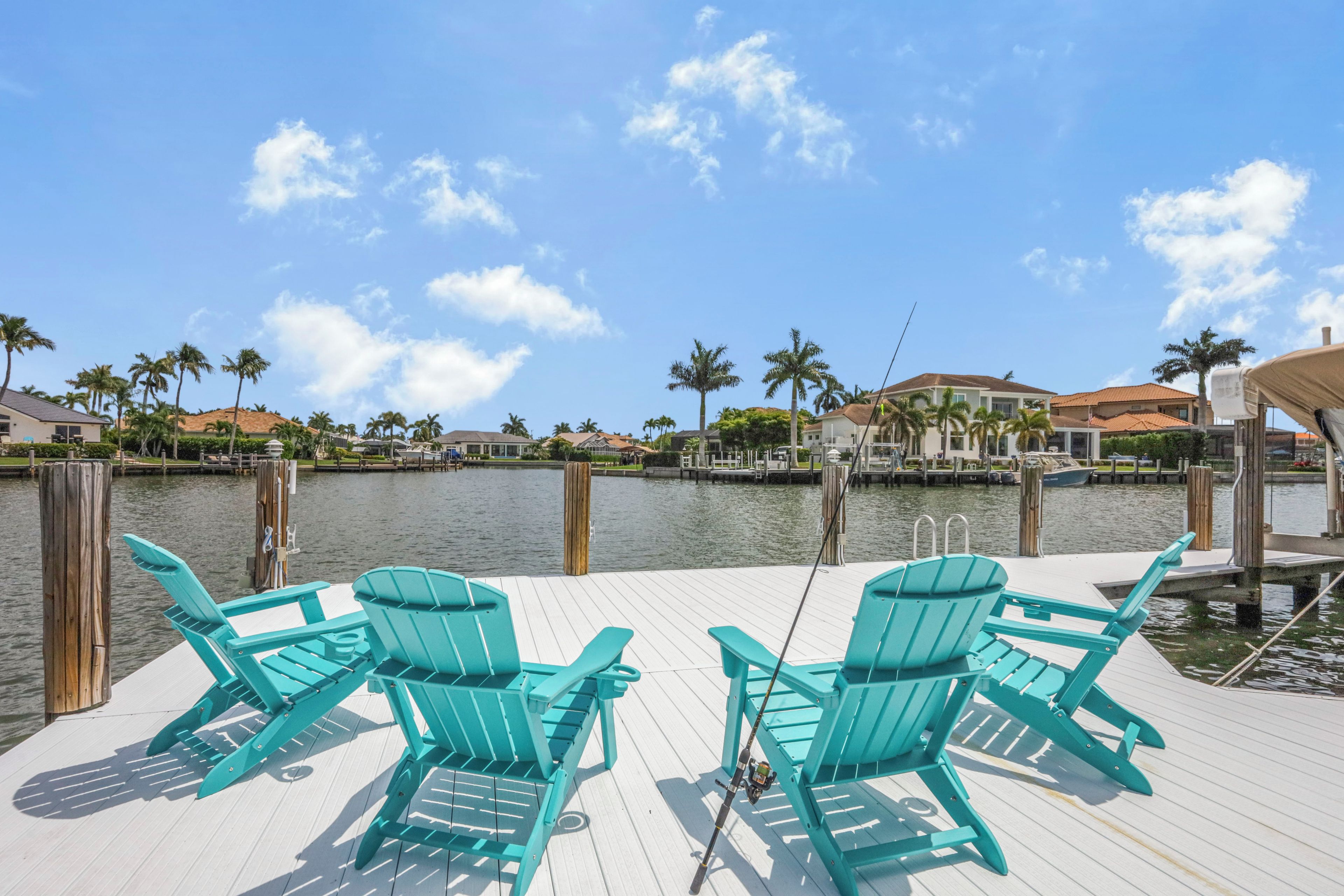 A dock with two chairs and a fishing rod, overlooking a calm body of water with houses and palm trees in the background.