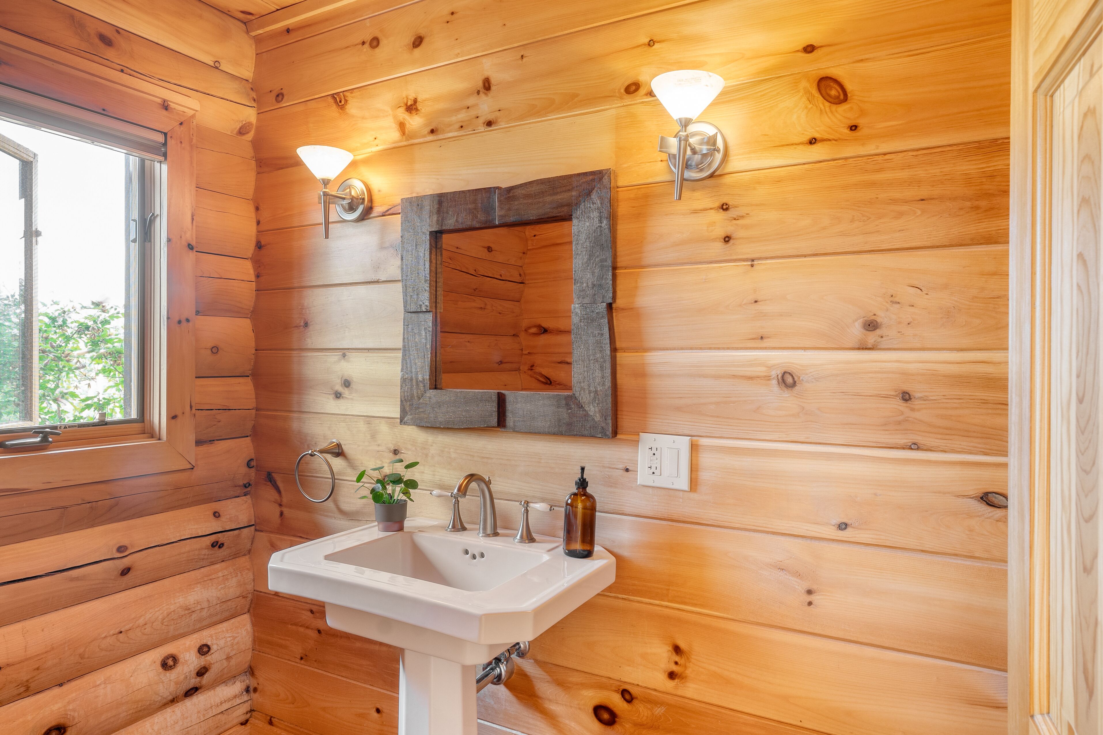 Un baño de cabaña de madera con un lavamanos blanco, dos luces empotradas en la pared, un espejo y una ventana.