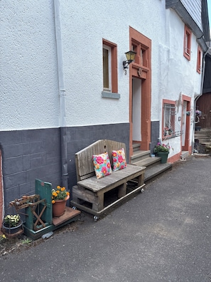 A wooden bench with floral cushions outside a building with a white and grey facade, a small step leading to the entrance, and a potted plant with yellow flowers.