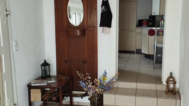 Een kamer met een houten deur, een kleine tafel met een lantaarn, een vaas met bloemen en een keuken op de achtergrond.
