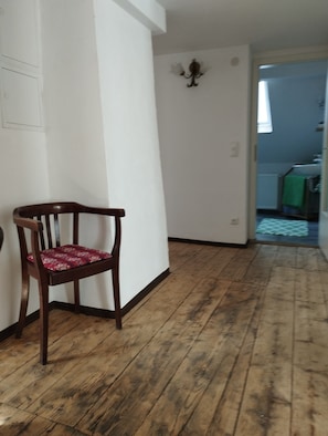 A hallway with wooden flooring, a wooden chair with a patterned cushion, and a wall-mounted light fixture.
