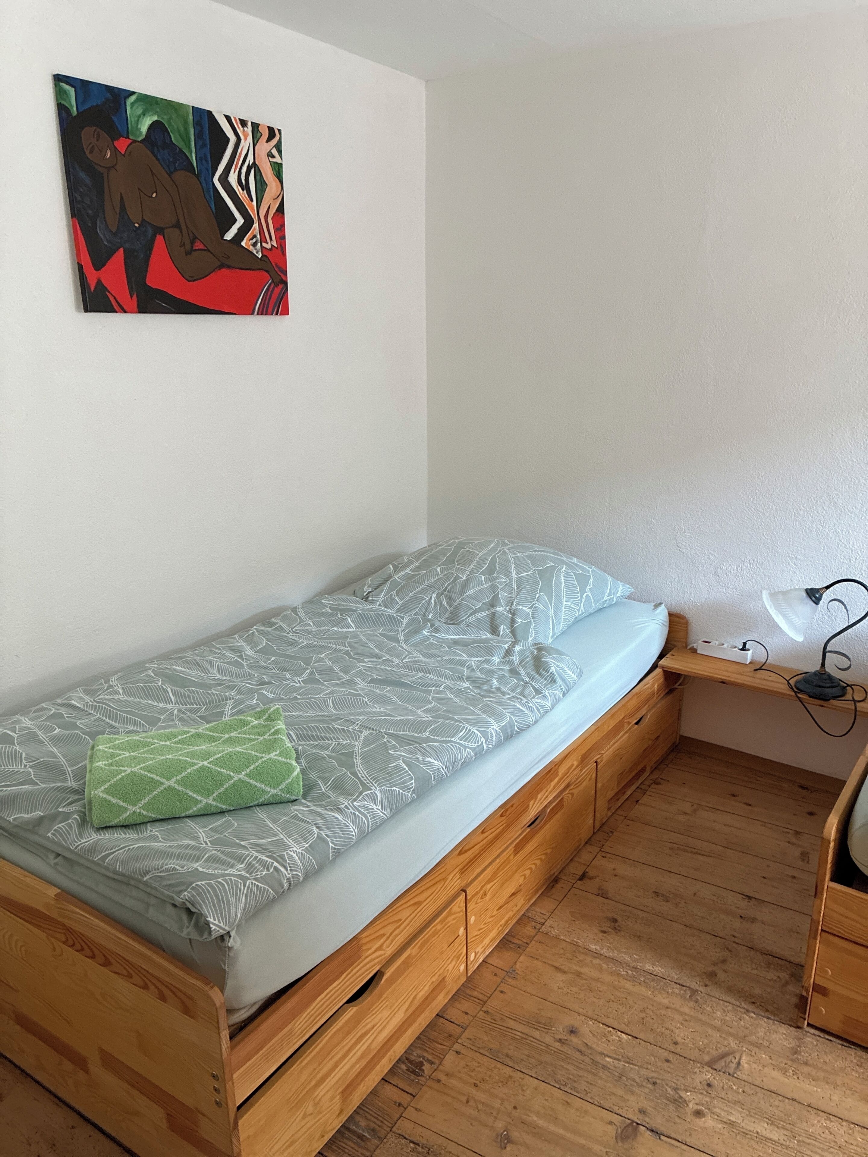 A small, single-bed room with a wooden bed frame, a patterned mattress, a green towel, a wall-mounted abstract painting, and a desk with a lamp.