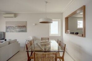 Un coin repas avec une table en verre, des chaises en osier, un luminaire suspendu, un tableau au mur et un téléviseur posé sur un canapé.