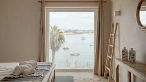 A bedroom with a large window, a bed, a bedside table with bottles, a mirror, and a view of the sea. - Apartment 'Cascais Bay Terrace IV' with Sea View & Wi-Fi (Cascais)