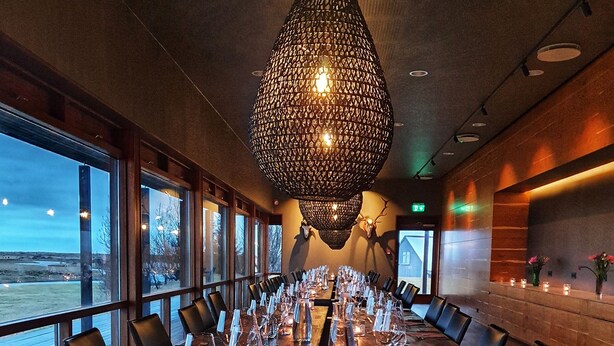 A long wooden table set for a meal with candles, glasses, and cutlery, in a room with large windows and a hanging light fixture. - West Ranga lodge (Rangárþing ytra)
