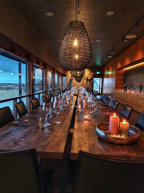 A long wooden table set for a meal with candles, glasses, and cutlery, in a room with large windows and a hanging light fixture.