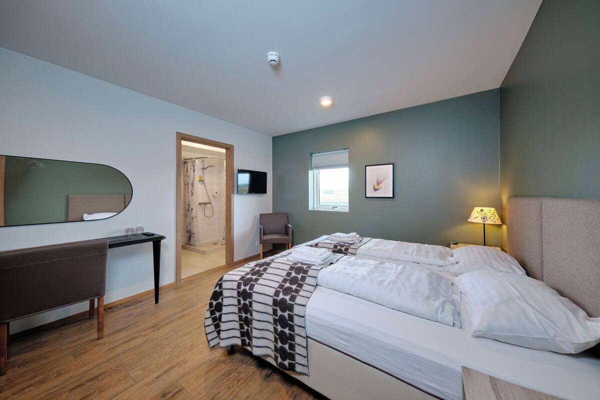 Quarto de hotel moderno com cama, escrivaninha, cadeira, chuveiro e televisão na parede.