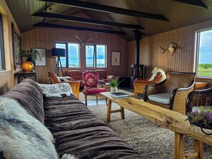 A cozy room with a long sofa, a wooden coffee table, and two chairs. There is a fireplace and a large window with a view. - West Ranga lodge (Rangárþing ytra)