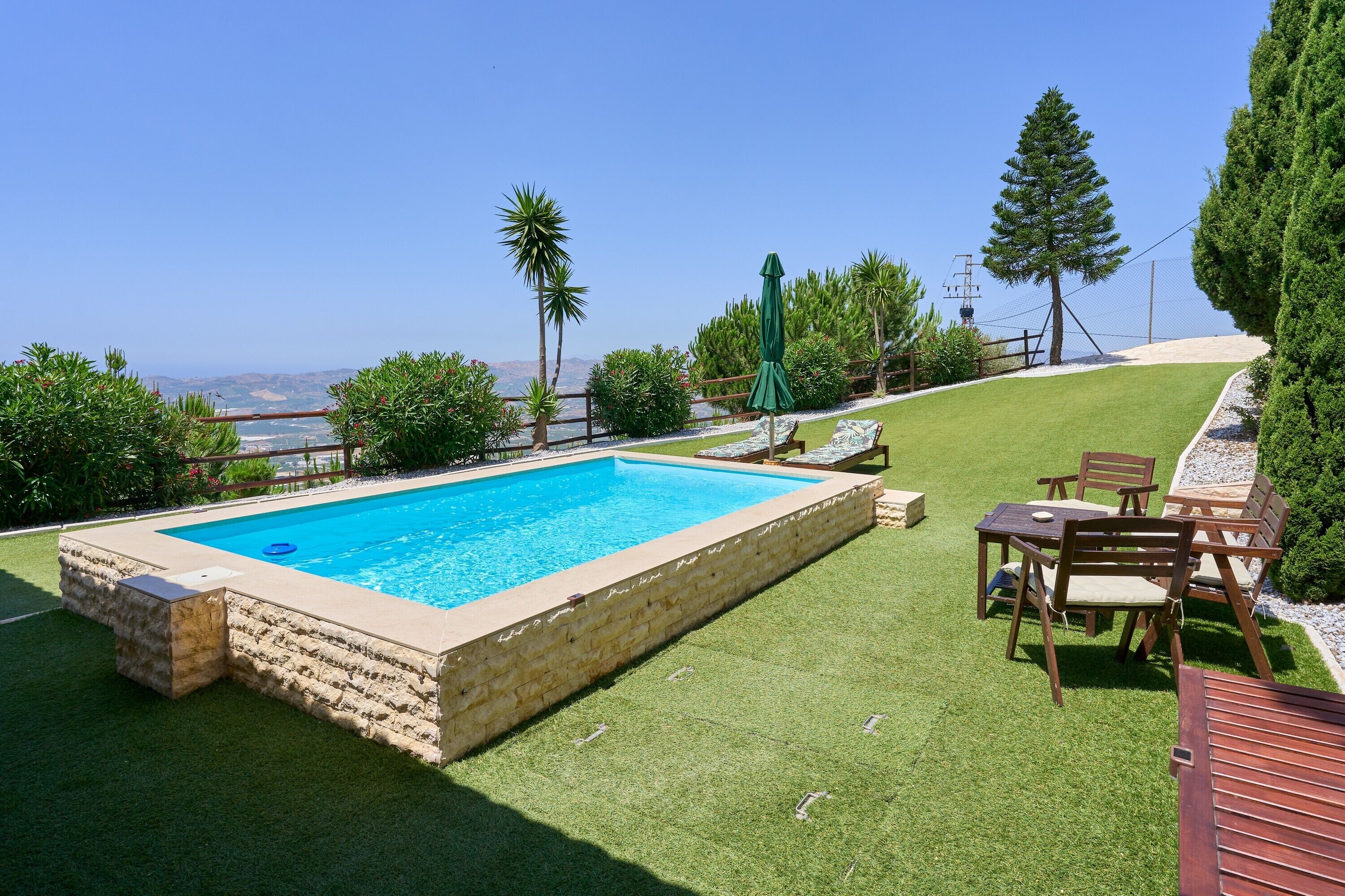 A rectangular swimming pool with a stone border, surrounded by artificial grass, a wooden deck, and a view of the ocean.