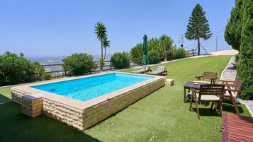 A rectangular swimming pool with a stone border, surrounded by artificial grass, a wooden deck, and a view of the ocean.
