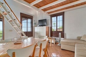 Un salon avec un escalier en bois, un téléviseur fixé au mur, une table à manger avec des chaises, un canapé et une fenêtre donnant sur un paysage verdoyant.