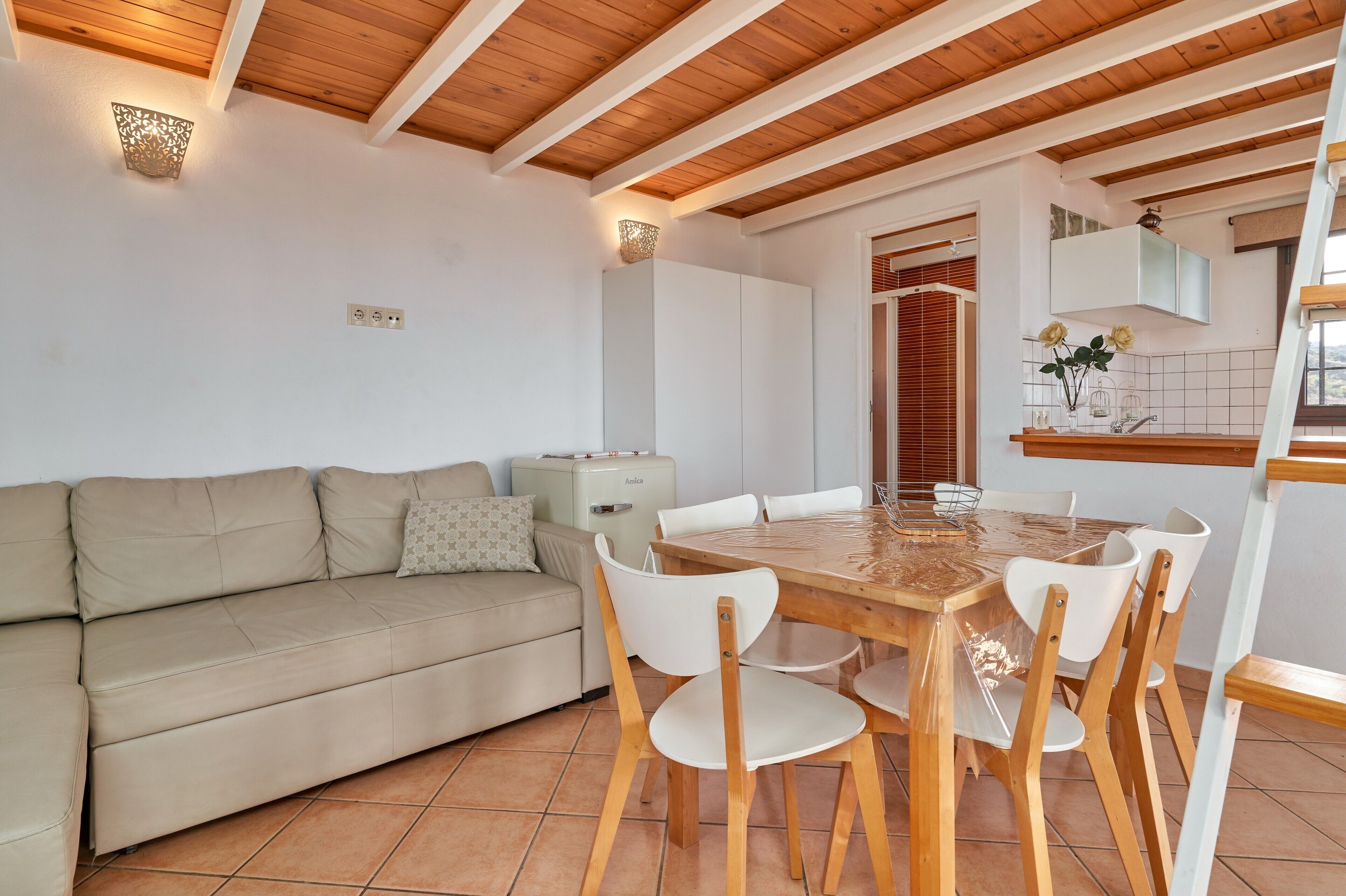 Une pièce avec un plafond en bois, un canapé beige, une table à manger en bois avec des chaises, un coin cuisine et une fenêtre avec des stores.
