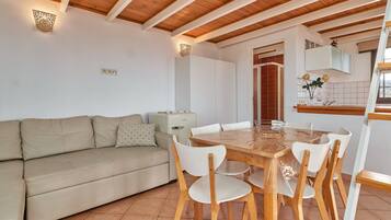 Une pièce avec un plafond en bois, un canapé beige, une table à manger en bois avec des chaises, un coin cuisine et une fenêtre avec des stores.