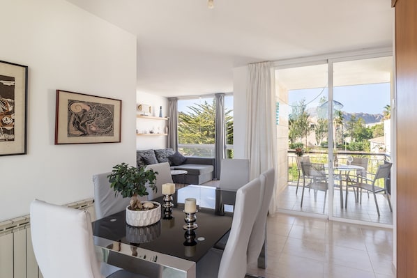 A modern living room with a glass dining table, white chairs, a sofa, and a balcony with a view of greenery and mountains.